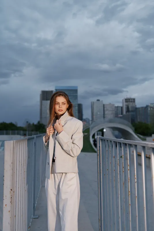 Woman in beige jacket and white pants stands confidently on urban bridge with cloudy sky and modern cityscape in background, fashion business style woman.