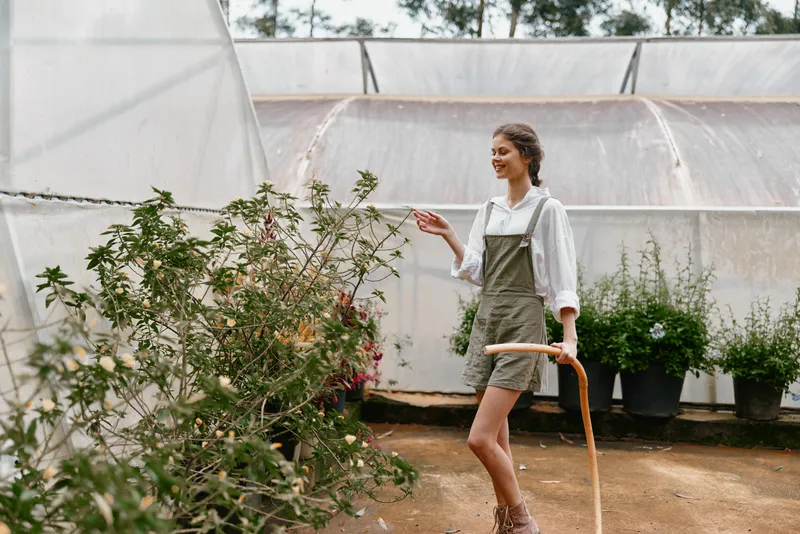 Young woman in casual green overalls gardening in a greenhouse, showcasing joy and connection to nature with vibrant plants around her