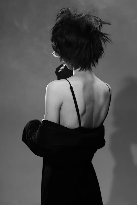 Black and white portrait of a woman with short hair and bare back wearing a strap dress and coat, artistic and moody atmosphere.