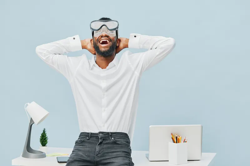 Happy young man in white shirt wearing protective goggles and smiling with hands behind head, standing at modern office desk isolated on light blue background. Workplace, casual, positive emotion, int