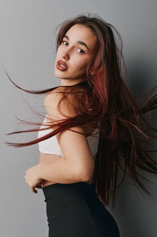 Young woman with long red hair posing in studio with hair flowing, wearing white top and black pants, looking over shoulder with natural makeup on gray background.