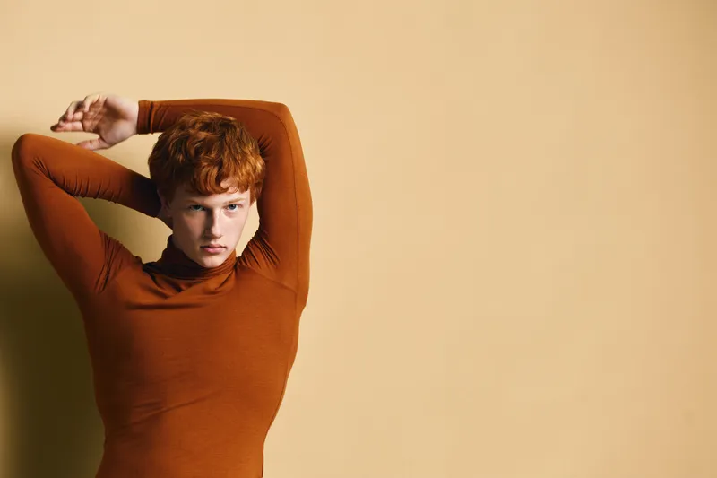 Young woman with red hair wearing brown sweater posing with arms raised against plain beige background in studio. Female model with short hair and serious expression in casual clothing.