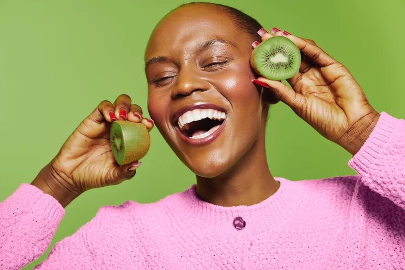 Happy black woman with dark skin holds fresh kiwi halves near face smiling with closed eyes on green background. Concept of healthy lifestyle, fruit, skincare, wellness, and natural beauty.