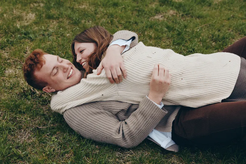 Romantic couple relaxing on grass, lying close and smiling, warm sweatered duo enjoying a carefree moment outdoors, intimate embrace, casual fashion, natural light, tranquil park setting, candid