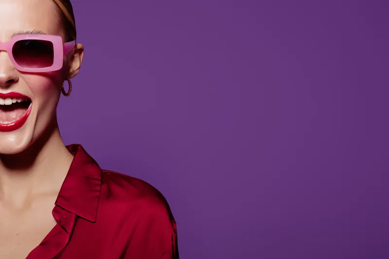 Happy woman with pink sunglasses and red lipstick smiles excitedly against purple background. Fashion concept with vibrant colors and joyful expression in studio lighting.