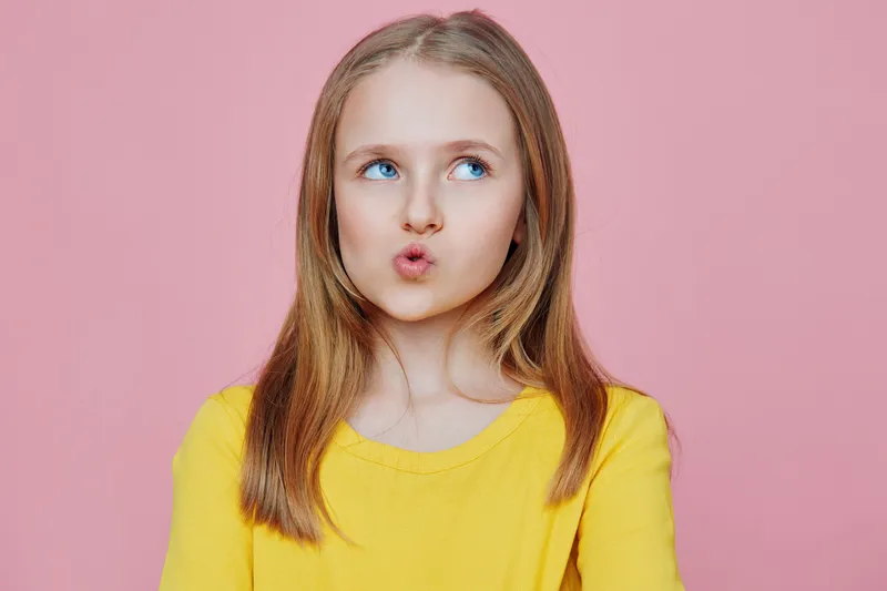 Young girl with long hair making a playful facial expression, wearing a bright yellow shirt against a pink background, symbolizing joy and innocence