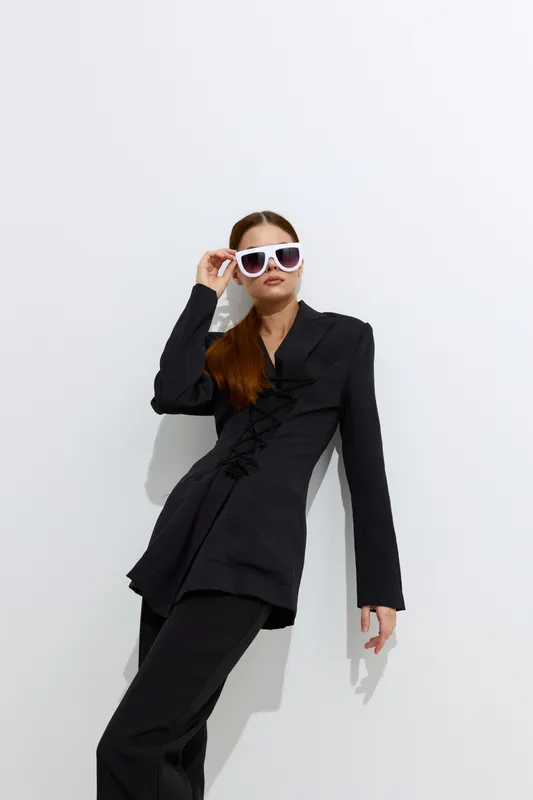 Fashion woman in black suit and white sunglasses poses confidently against white background, showcasing modern style and chic attitude with sleek hair and minimalist composition.