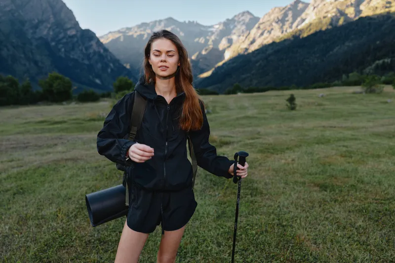 A confident outdoor explorer stands in a wide valley with towering mountains behind, wearing a black jacket and shorts, carrying a camera bag and trekking pole.