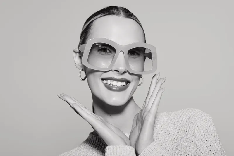 Smiling white woman with fair skin wearing large square sunglasses and a knitted sweater poses with hands near face. Studio portrait on light background. Concept of fashion and eyewear.