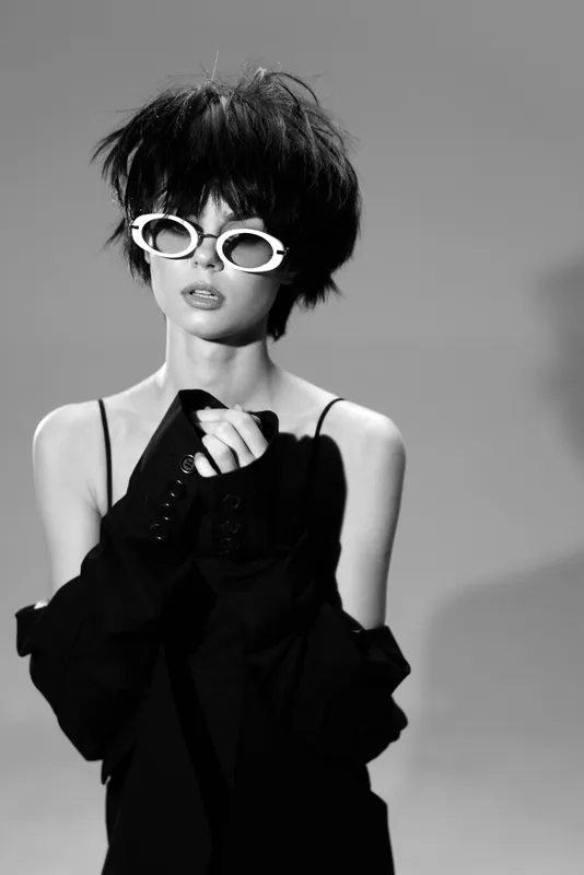 Black and white portrait of a stylish woman wearing white sunglasses and dark clothing, expressing contemporary fashion and edgy attitude.