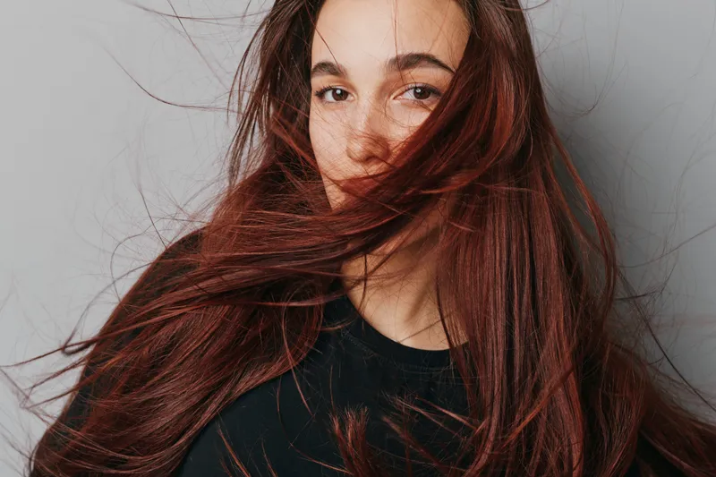 Young woman with long red hair flowing across her face wearing black top against neutral background. Portrait with natural expression, casual style and loose hair movement in studio setting.