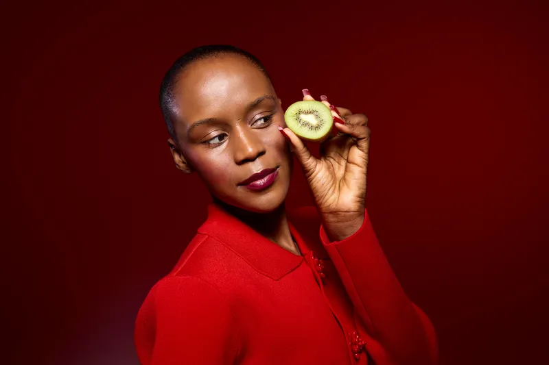 Elegant black woman with dark skin holding a sliced kiwi near her face, wearing a red sweater against a deep red background. Concept of healthy lifestyle, beauty, and fresh fruit.