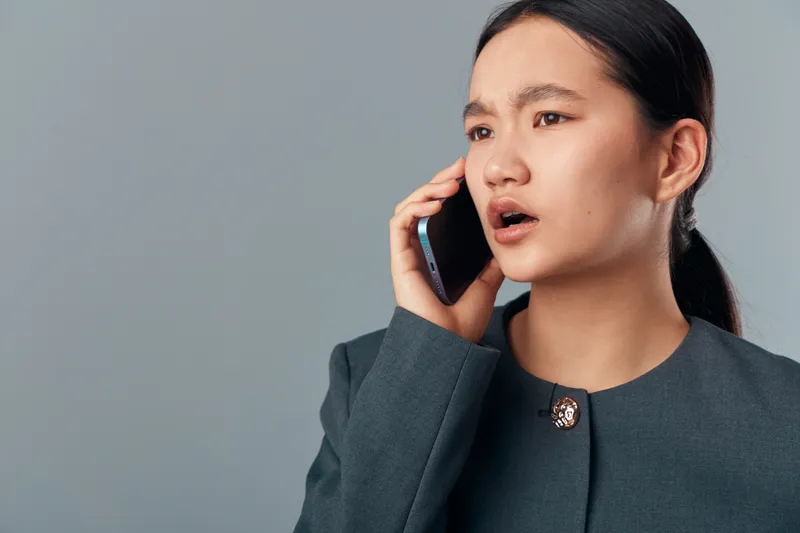 Woman with serious expression talking on smartphone against grey background. Businesswoman in formal jacket using mobile phone for important conversation or communication.