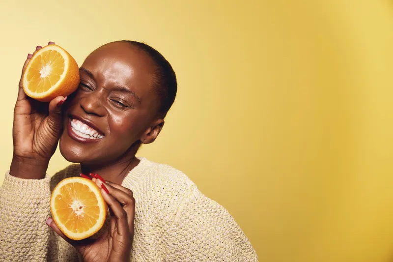 Happy African American woman with dark skin holds two orange halves near face smiling with closed eyes on yellow background. Concept of healthy lifestyle, vitamin, and natural beauty.