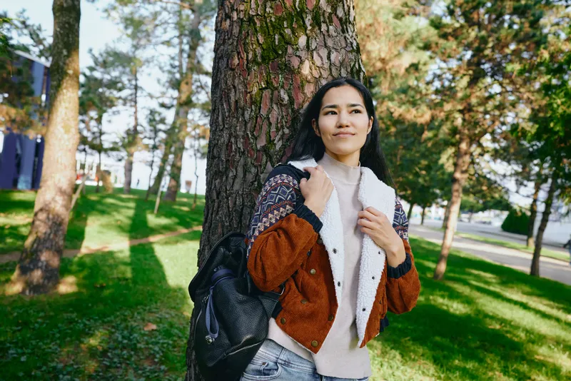 Woman leaning against a sturdy tree in a sunlit park, wearing a brown jacket and light scarf, carrying a backpack, enjoying a casual outdoor moment amid green grass and autumn colors.