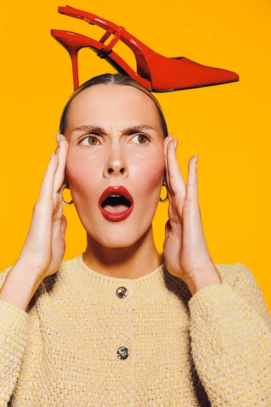 Surprised woman with red lipstick holds head with hands and balances red high heel shoe on her head. Bright yellow background creates vibrant and playful fashion concept.
