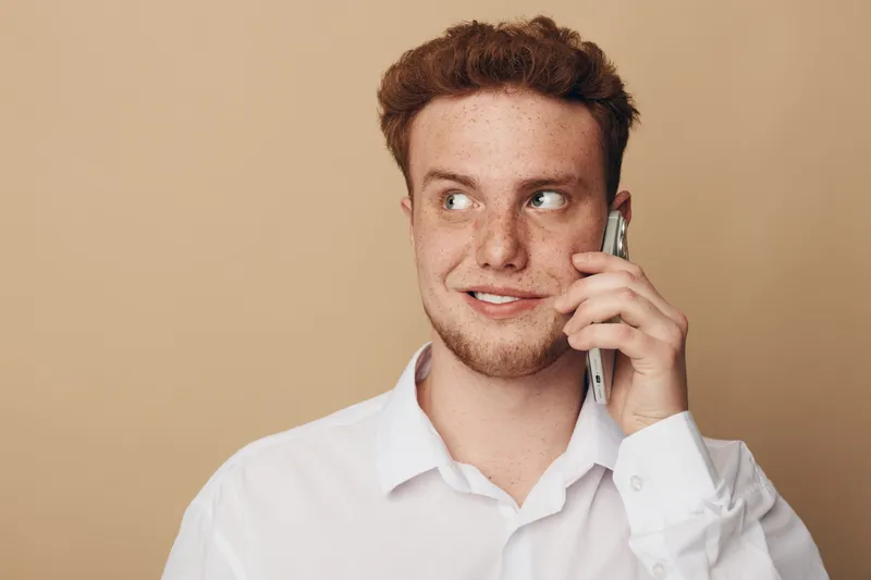 Young man with red hair and freckles wearing white shirt talks on smartphone looking aside with slight smile. Neutral beige background. Concept of communication, technology, and casual lifestyle.