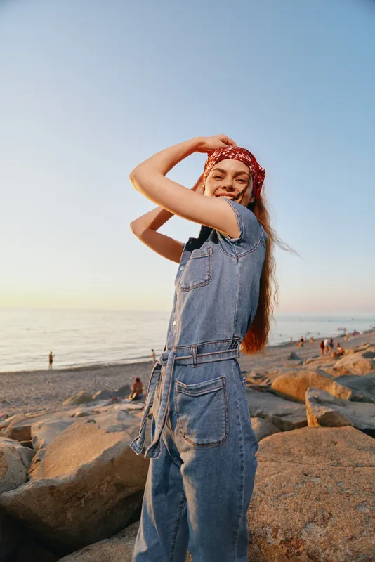 smiling woman in denim jumpsuit and red bandana enjoying sunset at rocky beach with calm sea and warm light