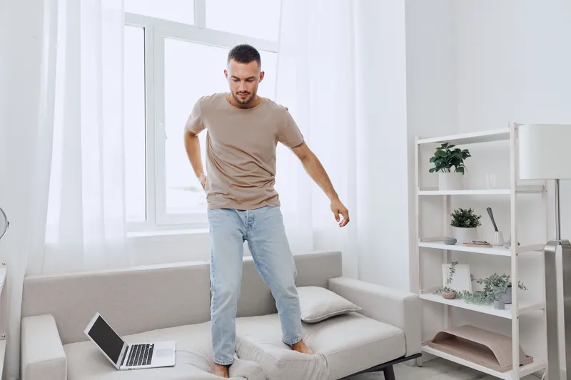 A young man confidently balancing on a couch while using a laptop, showcasing modern remote work dynamics and home comfort in a bright, minimalist setting.