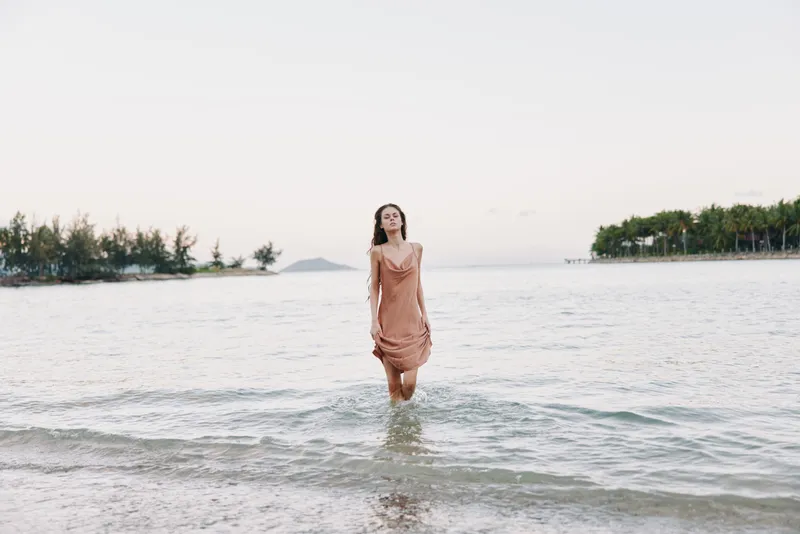 A woman in a soft peach dress stands in shallow water near the shoreline, creating a serene, graceful scene with gentle waves and a peaceful coastal mood.