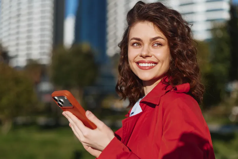 A smiling woman in a red jacket holds a smartphone outdoors in a sunny urban park, conveying warmth, confidence, and everyday technology use in a casual moment.