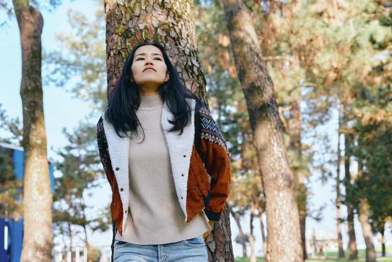 Woman outdoor forest trees jacket sweater casual confident looking upward with calm, natural sunlight and peaceful mood in a forest setting, airy ambience and gentle breeze