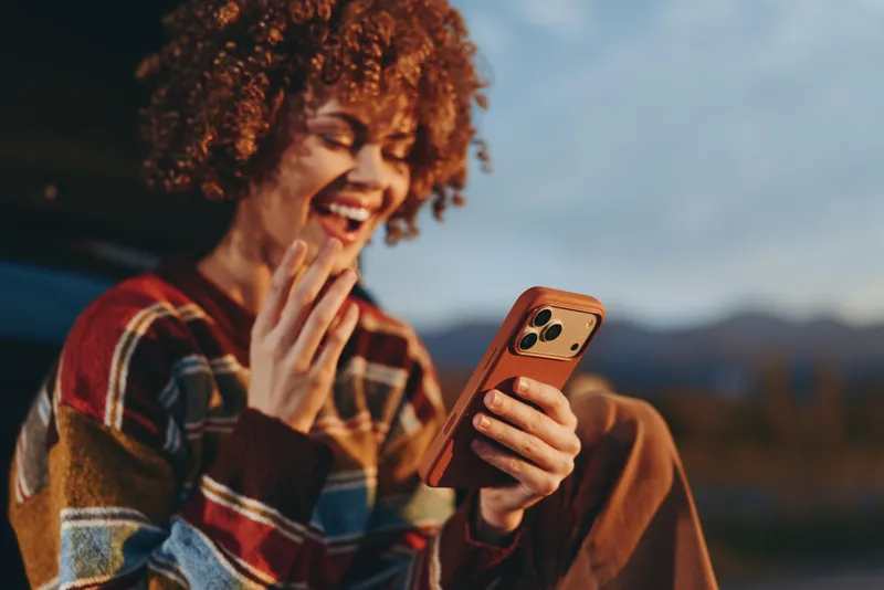 Woman with curly hair smiles outdoors, holding smartphone with communication gesture, casual lifestyle, mountain background, warm golden light, shallow depth of field
