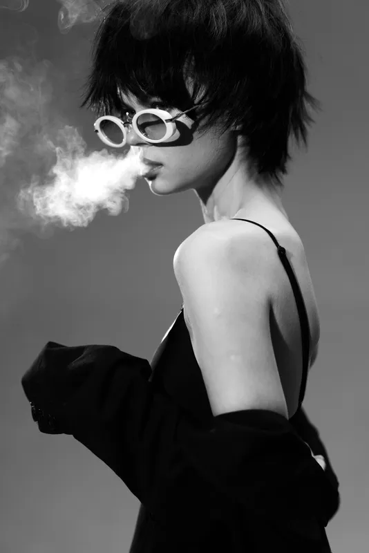 Black and white portrait of a young woman in sunglasses exhaling smoke with an edgy and mysterious style