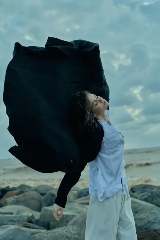 Woman on a rocky beach with a flowing cape, wind catching fabric against a moody sky, creating dramatic movement and a fashion driven outdoor scene.