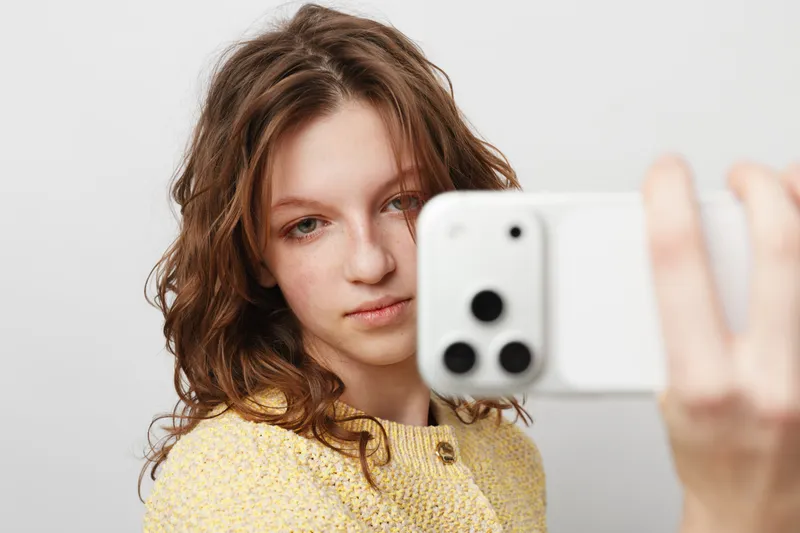 Young woman with natural makeup and wavy hair takes a selfie with a smartphone on a white background. Concept of casual lifestyle, technology, and self-portrait photography.