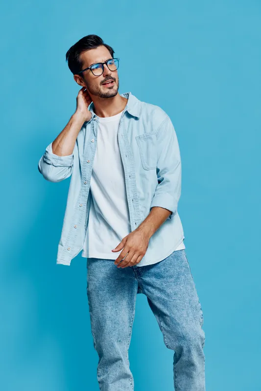 Stylish young man with glasses posing confidently in a light blue shirt and denim jeans against a solid blue background. Fashion concept and modern lifestyle.