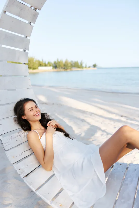 A smiling woman in a white dress lounges on a curved wooden seaside chair, sunlit beach, calm blue water, and distant trees creating a serene summer scene.