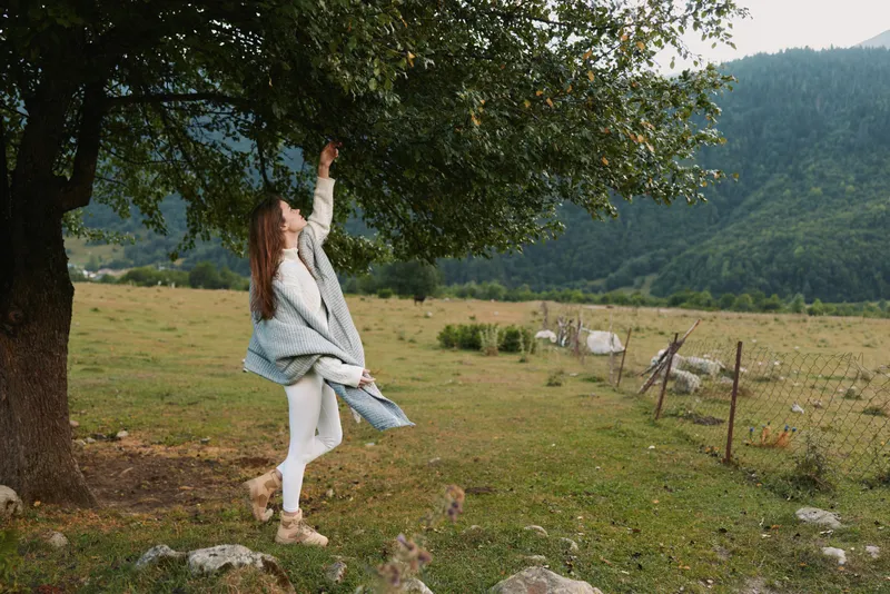 Cheerful woman outdoors reaching up to a tree branch, casual lifestyle moment, sunlit meadow, gentle breeze, natural colors, serene countryside background