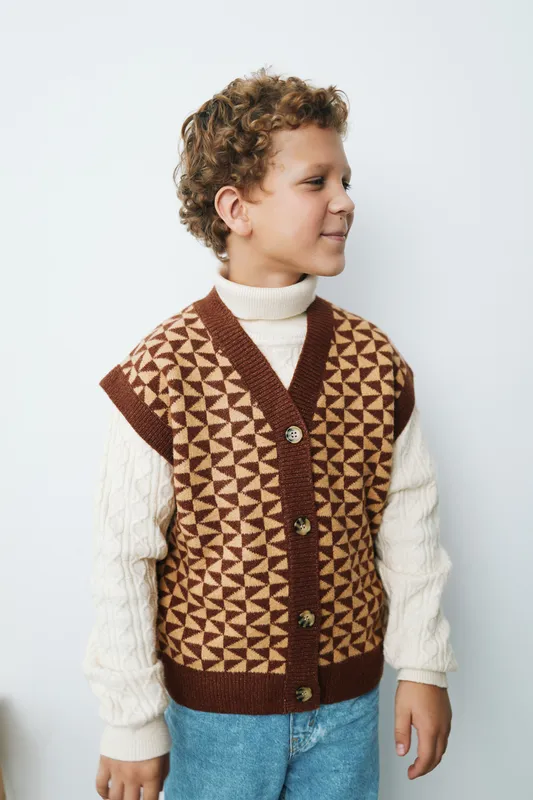 Young boy wearing a geometric patterned knit vest over a cream turtleneck, facing right, standing against a light background, showcasing cozy autumn style and playful innocence.