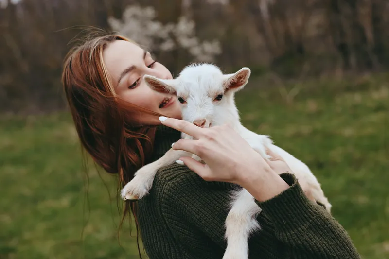 Smiling woman with long hair holding a white goat kid in a green field. Close-up on a natural backdrop. Lifestyle moment celebrating care, connection, and gentle affection between human and animal.