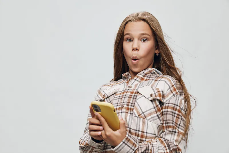 Girl with long hair and plaid shirt holding yellow smartphone, showing surprised emotion on face with wide eyes and open mouth on gray background.