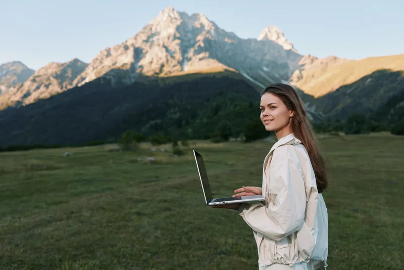 A thoughtful woman stands outdoors with a laptop in a scenic mountainous landscape. She looks focused yet relaxed, creating a calm and productive setting for remote work.