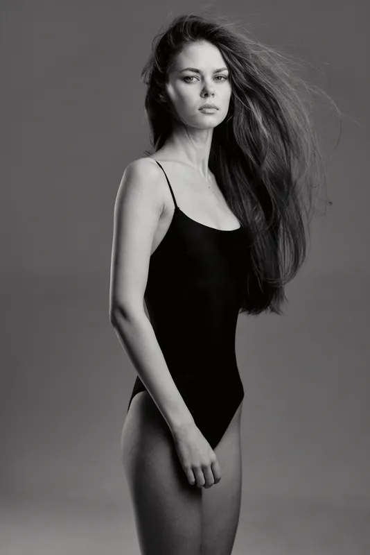 Black and white portrait of a young woman in a black bodysuit with long hair and confident expression.