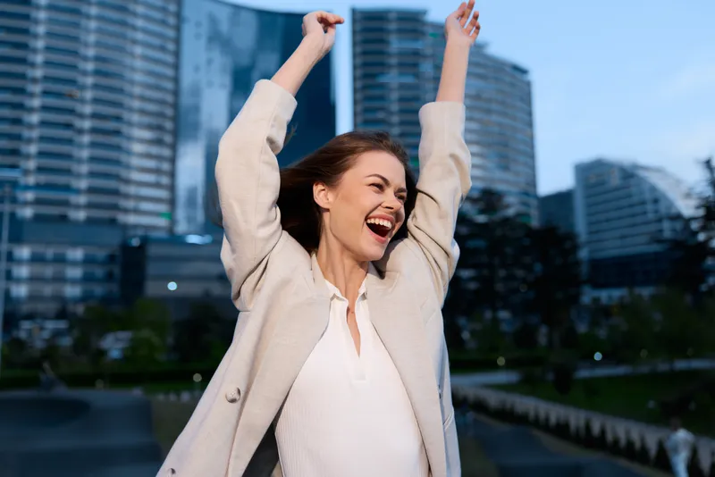 Confident woman celebrating success in a city park, dressed in a stylish light jacket and top, with a joyful expression against a modern urban backdrop.
