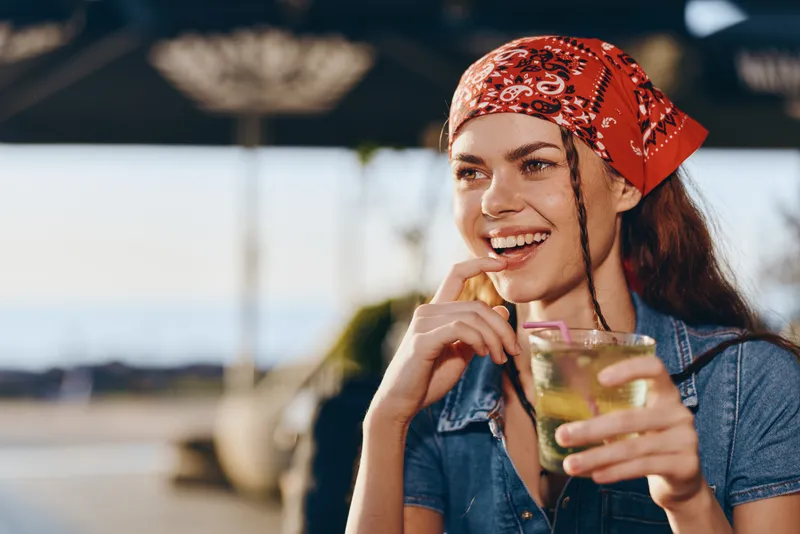 Lifestyle woman in modern boho-western denim street style smiling and holding a refreshing drink with a film color tone and warm outdoor background.
