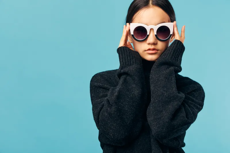 Woman wearing white sunglasses and black sweater posing against blue background for fashion portrait style, casual look with confident expression, modern clothing and trendy eyewear.