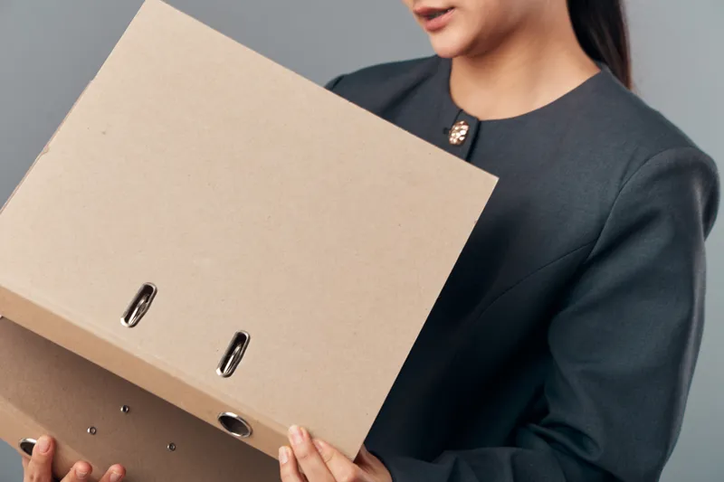 Woman holding cardboard file folder with metal rings, wearing business suit, organizing documents for office work. Closeup of hands and folder on neutral background for paperwork and administration.