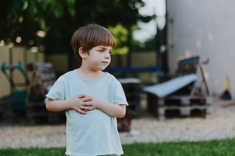 Thoughtful boy standing outdoors, wearing a light blue shirt, surrounded by greenery and wooden pallets, reflecting a sense of curiosity and innocence.