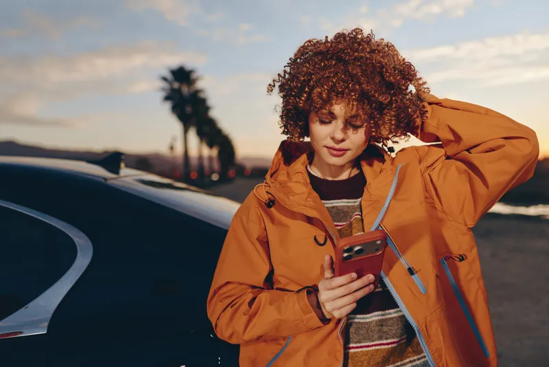 Woman lifestyle outdoors near car, holding smartphone and looking down, curly hair and tan jacket, sunset sky over road, hand on head, travel mood, evening breeze