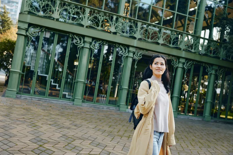 Woman in beige coat with sweater and jeans stands on a city street beside ornate green railings, casual fashion, outdoor scene, friendly smile, daytime urban ambiance