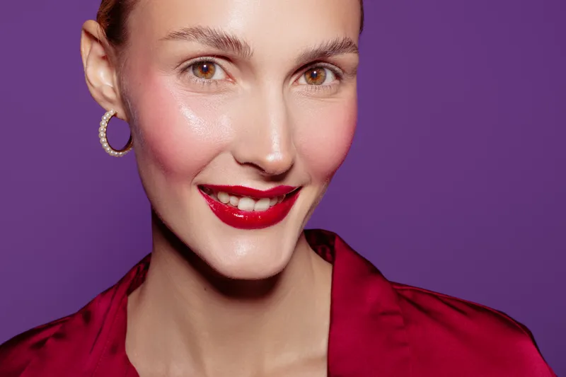 Close-up of smiling woman with red lipstick and hoop earrings wearing red satin blouse on purple background. Concept of beauty, makeup, fashion, and confident expression.