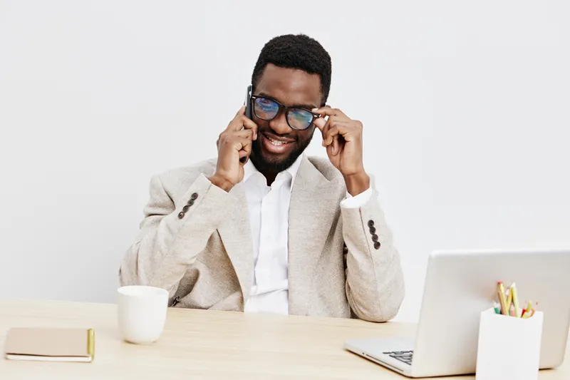 Smiling man with dark skin, curly black hair, wearing glasses and beige blazer, working at desk with laptop and cup on bright white background, business casual style, professional, relaxed, modern off