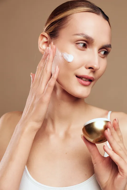 Young woman with clear skin applying white face cream on her cheek, holding a golden jar, with a neutral brown background, concept of skincare, beauty, and self-care routine.