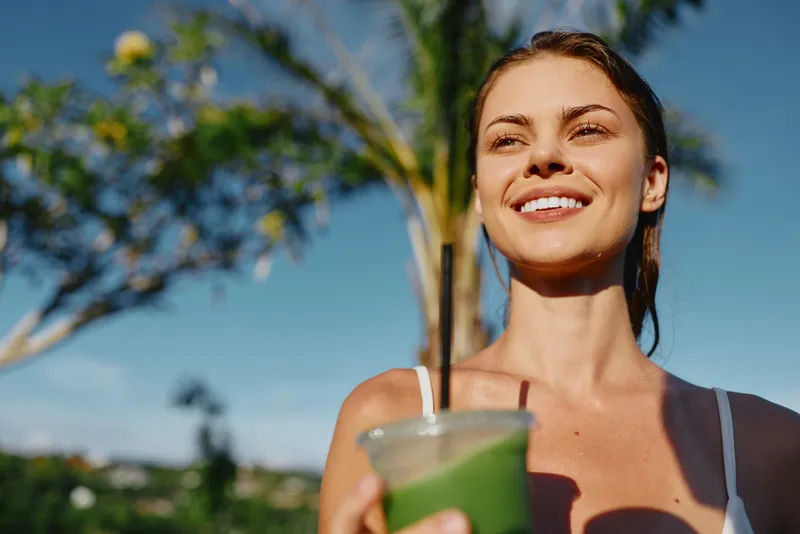 Cheerful woman enjoys a refreshing green drink outdoors, smiling under bright sun, casual summer vibe, sunlit skin, relaxed portrait, vibrant garden backdrop, lifestyle moment captured during leisure