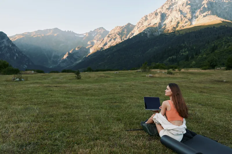 A woman sits on a grassy meadow with a laptop in a remote mountain setting. She appears focused and calm, enjoying a peaceful, productive outdoor workspace with expansive scenery and blue sky above.